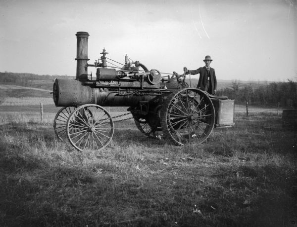 Ed Linnell stands on a steam tractor. Steam drives the horizontal piston, just behind the smokestack which runs the flywheel, on opposite side, which spun a long belt to operate implements such as a threshing machine. The tractor faces to left, Ed stands with his hand on the steering wheel, facing viewer. The two long chains to the front wheels were used to change direction of the wheels.