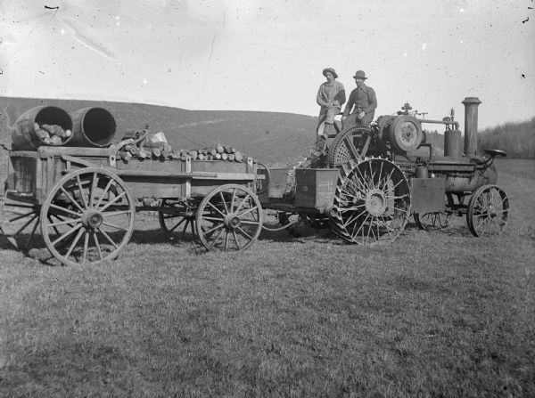 Two men sit atop a steam tractor that is pulling logs.