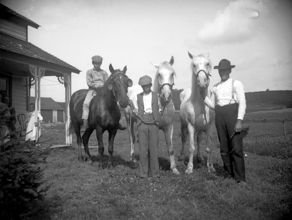Two men display three horses in front of a house. A boy sits on one of the horses, and a man, woman, and girl stand on the house porch.	