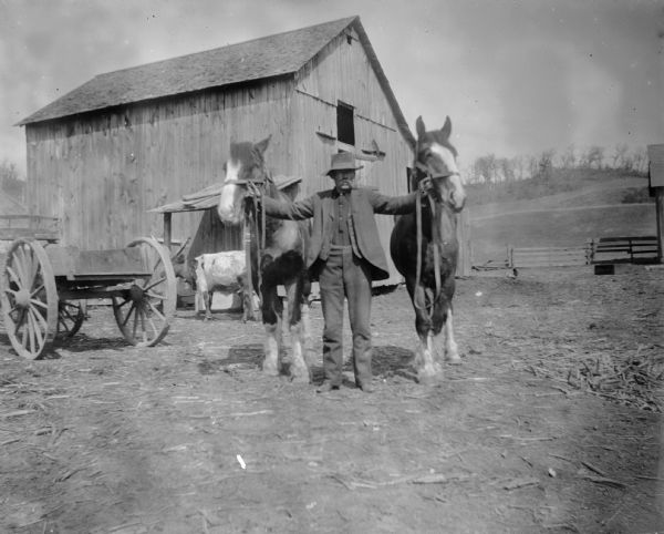A man holds two horses by their reins in front of a barn. A wagon is on the left.