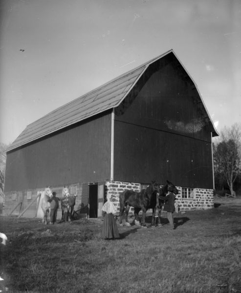 Two men each hold a pair of horses outside of a stone foundation barn as a woman observes.