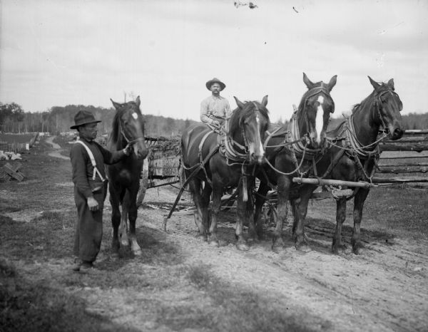 A farmer drives a team of three horses hitched to a manure spreader. Another man stands next to the team, holding an additional horse by its reins.