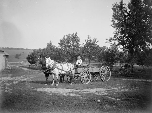 Two horses pull a farmer's wagon. The corner of a building is on the left, and trees are in the background. A hill is in the distance.