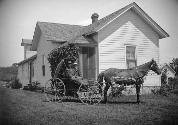 Two men sit in a buggy pulled by a horse wearing a fly-net. The buggy pauses in the yard in front of a frame house. 	