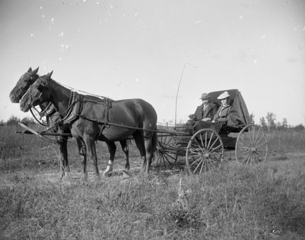 A man and woman sit in a buggy that is pulled by a two-horse team.