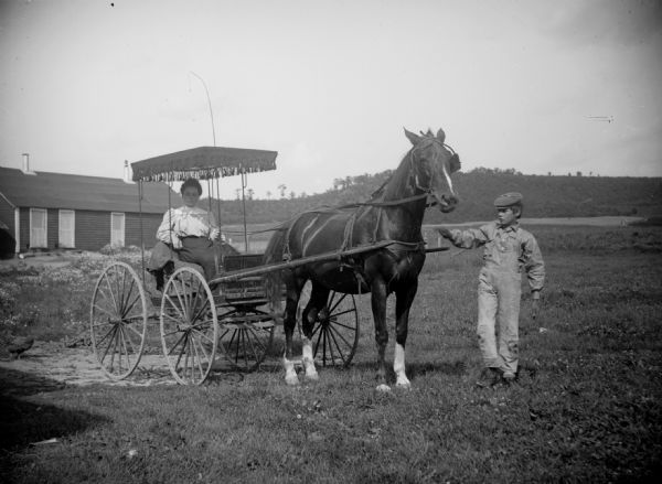 A boy wearing a cap is leading a horse that is pulling a woman sitting in a buggy. A building is in the background on the left.