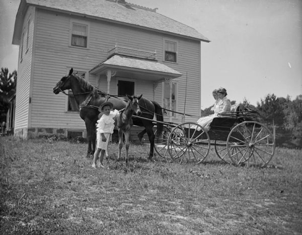 A young boy is standing with a colt near two women who are sitting in a horse-drawn carriage in front of a house.