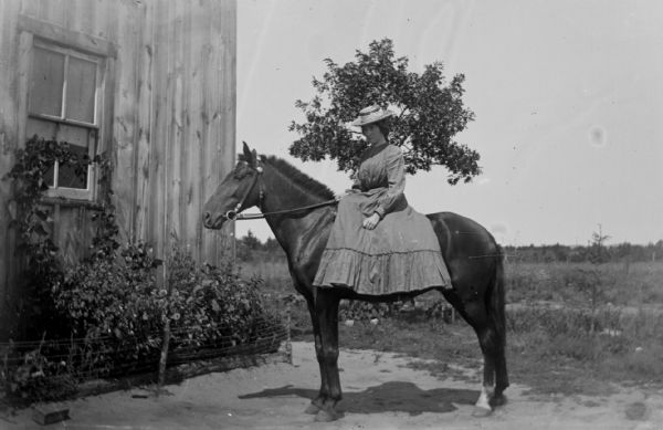A woman sits side-saddle on a horse in front of a farm building.