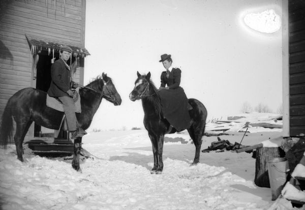 A man and woman pose on separate horses in a snow-covered farmyard.