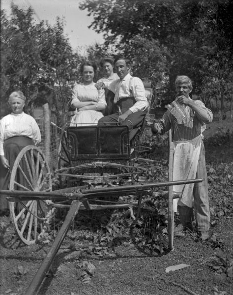 Two women and a man sit in a wagon, while another man and woman stand on the outside of the wagon.