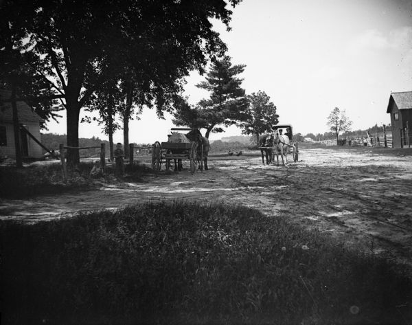 Two carriages are on a dirt road of a farm under trees. Children are sitting and standing in the yard on the left.