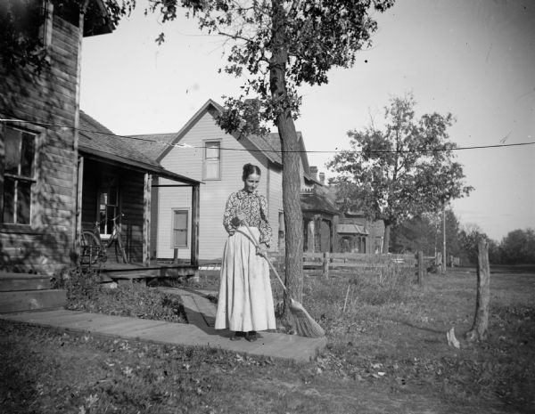A woman (possibly a member of the Weller family) sweeps a board sidewalk with a broom. On the left is the Weller home, and next to it is the Carl Lund house. This is a view of Madison Street, looking south from the corner of Third Street, in "The Grove. "	