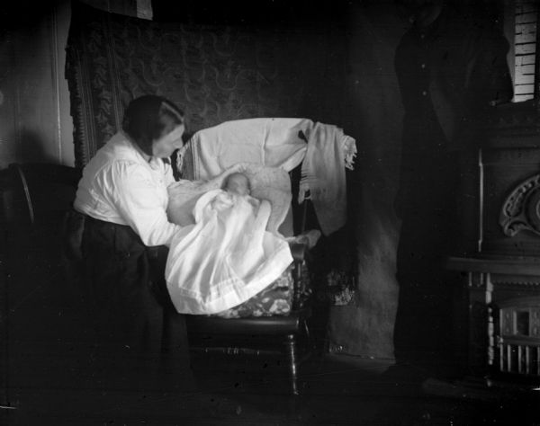 A woman in a light top and dark skirt tending to a baby propped on a chair wearing a long white gown. A parlor stove is on the right, and tapestries are hanging on the wall in the background.