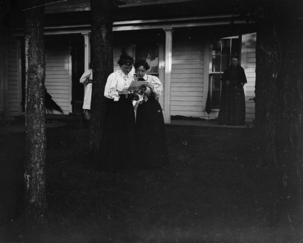 Two women stand in front of a house and jointly read a letter.