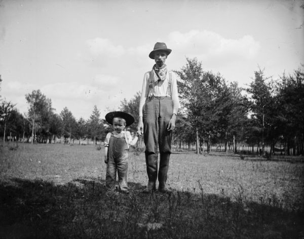 A farmer and a small boy, possibly a son, stand outdoors holding hands.