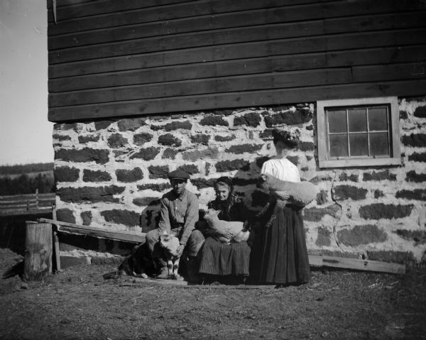 Two women and a man are posed next to a barn,each holding a lamb.