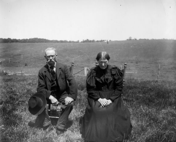 An elderly couple sits on chairs in a field. The woman's eyes are downcast.