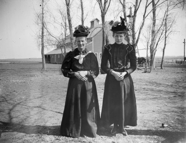 Two women pose standing with their hands folded, in a farmyard.