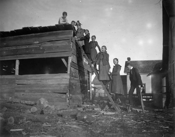 A group of seven children climb a ladder up to a wooden building while a man stands on the ground.