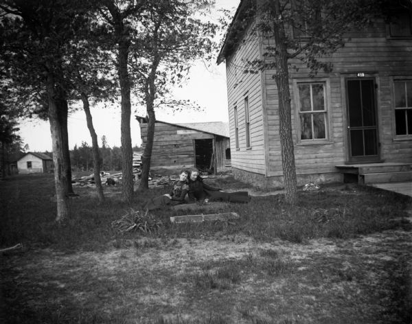Two women sit back to back in a yard near a farmhouse.