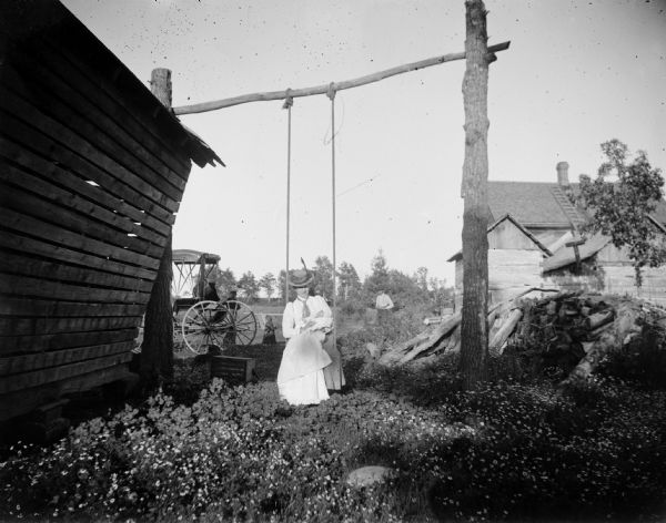 A woman sits on a homemade swing holding a baby. In the background,two men stand and watch another child.