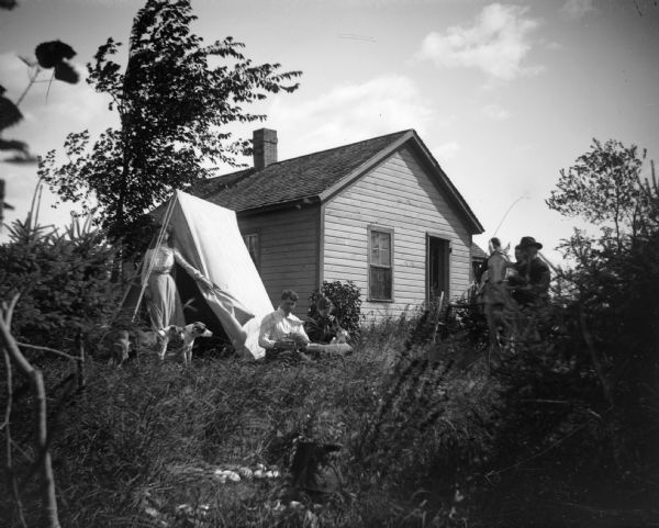 Several individuals, two dogs, and two cats gather around a tent set up near a building, while other individuals gather around a horse on the right.