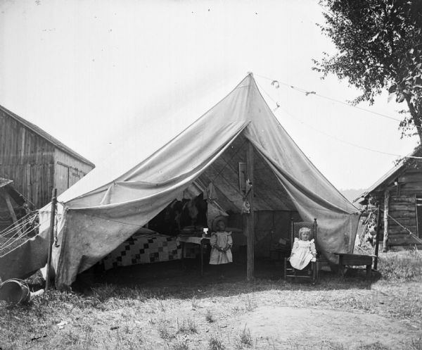 Two girls pose in an open tent that is pitched in a farmyard. Inside the tent there is a bed, clothesline and a table covered with books.