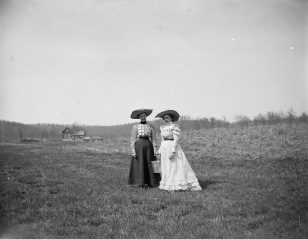 Two women wearing large hats stand in a field carrying a pail. Farm buildings are in the far background.