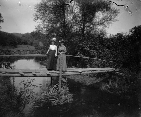Two women wearing fashionable hats stand on a footbridge over a stream.