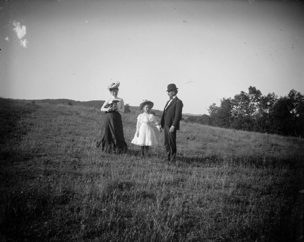 A woman, man and girl, possibly a family, stand in a field. The woman is holding a dog, and the man and girl are holding hands.	