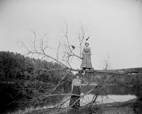 Two women stand on or near a fallen tree next to a river.	