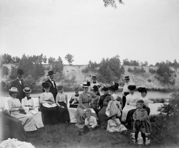 A group of men, women and children gather on the bank of a river.
