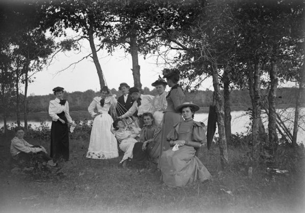 A group of men, women, and children relax in and around a hammock that is near a river.
