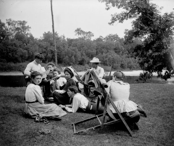 A group of men and women gather around a guitar player on the bank of a river.