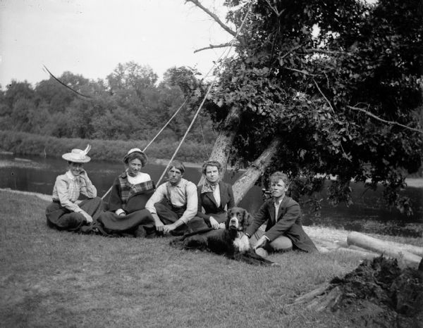 Three women, two men and a dog sit near a river. The men are smoking pipes and the woman second from the right may possibly be Edna Bright.