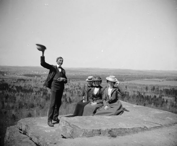 Three women and a man pose  at the top of a rock formation overlooking the flat fields and forest below. The three women are sitting while the man stands waving his hat.