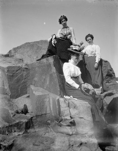 Three women sit on rocks holding their hats and gloves. The woman on the left and seated highest is probably Grace Jones Meck.	