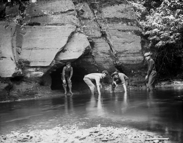 Four young men wade in the water at the base of a rock, one holding a fishing pole. The young men could possibly be the Taylor boys catching minnows in Perry Creek.