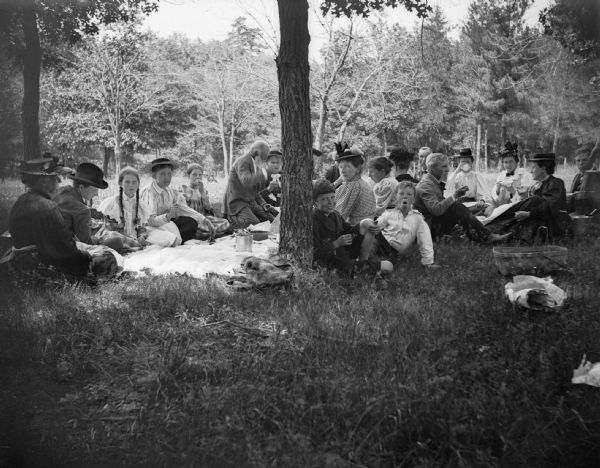 A large group of men, women, and children gather for a picnic. Between the two boys in the foreground, and wearing a small hat, is Mrs. Charles J. Van Schaick.	