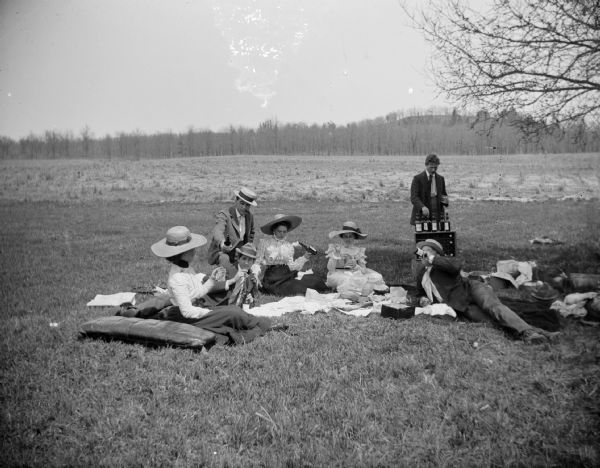 A group of men and women gather for a picnic and drink from glass bottles.