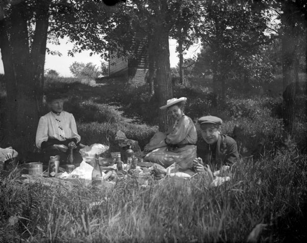 Two women and a man sit on the ground for a picnic amidst the trees. From from left to right; Mrs. Charles J. Van Schaick, Ewell(?), and either Harold or Roy Van Schaick.	