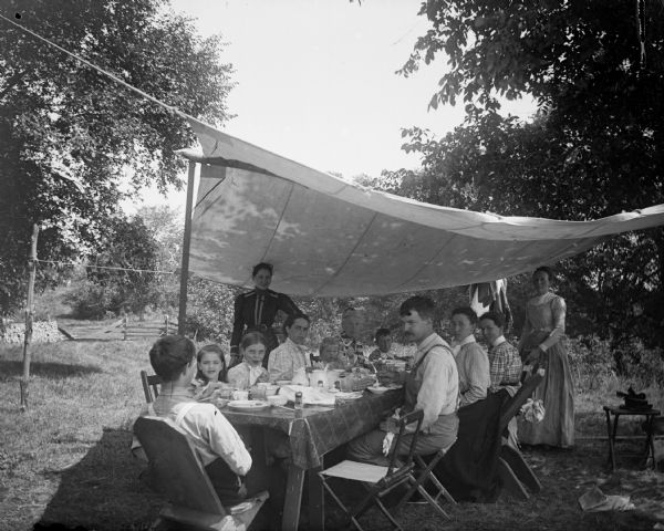 A large group sits at a long table having a picnic under an awning. A woodpile and a wooden fence are visible in the background.	