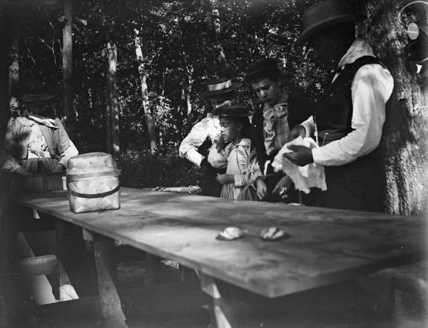 Three women, a girl and a man put out a meal on a picnic table in a wooded area.