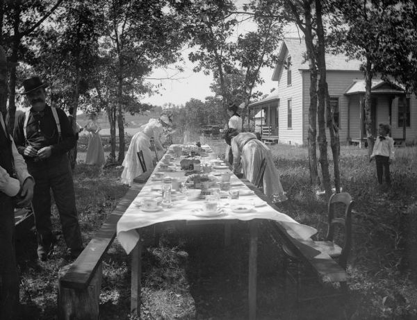 Three women set a long picnic table while two men and a young boy stand nearby.