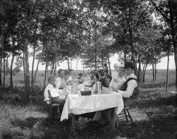 A large picnicking group sits at a long table among a grove of trees.