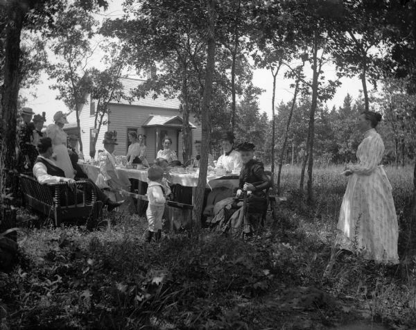 A large group of men, women and children gather for a picnic in a grove of trees near a frame house.