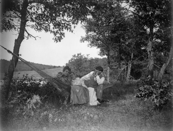 Two women and a man resting in a hammock that is tied between two trees.