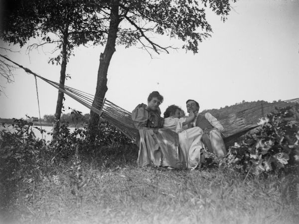 Two women sitting in a hammock with a man. One woman is straightening the man's bow tie.