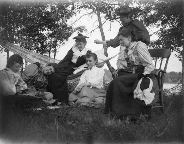 A group of women relax in or near a hammock between two trees. Identified from right to left: Flora LeClair (sitting), Lottie Bright (standing), Neddie Erickson, Julia Orrnsby, with the last two women unidentified.