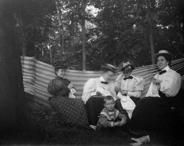 Four women sit in or near a hammock and attend to their embroidery while a young boy sits at their feet.